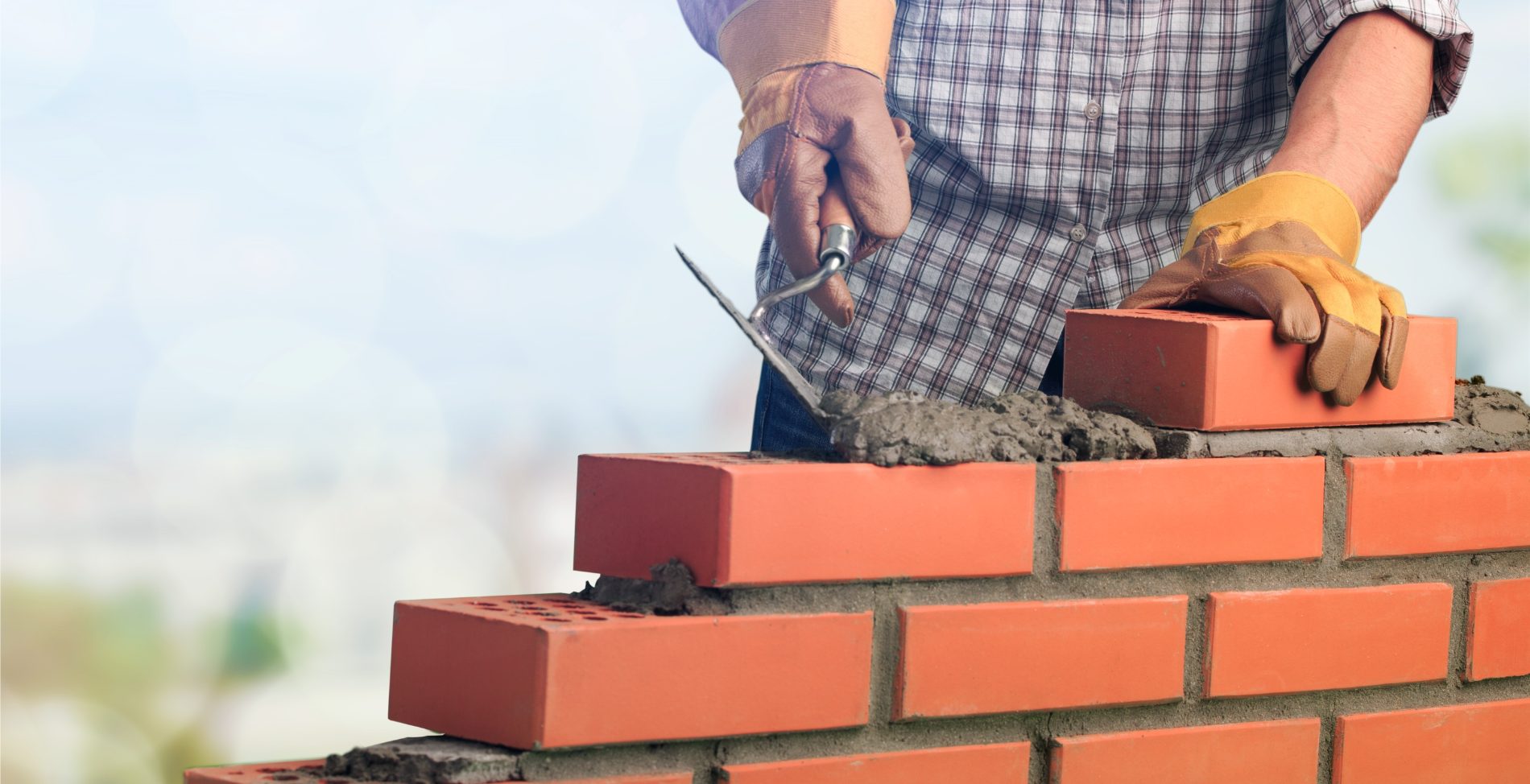 Mason in plaid shirt and yellow gloves laying red bricks with mortar and trowel