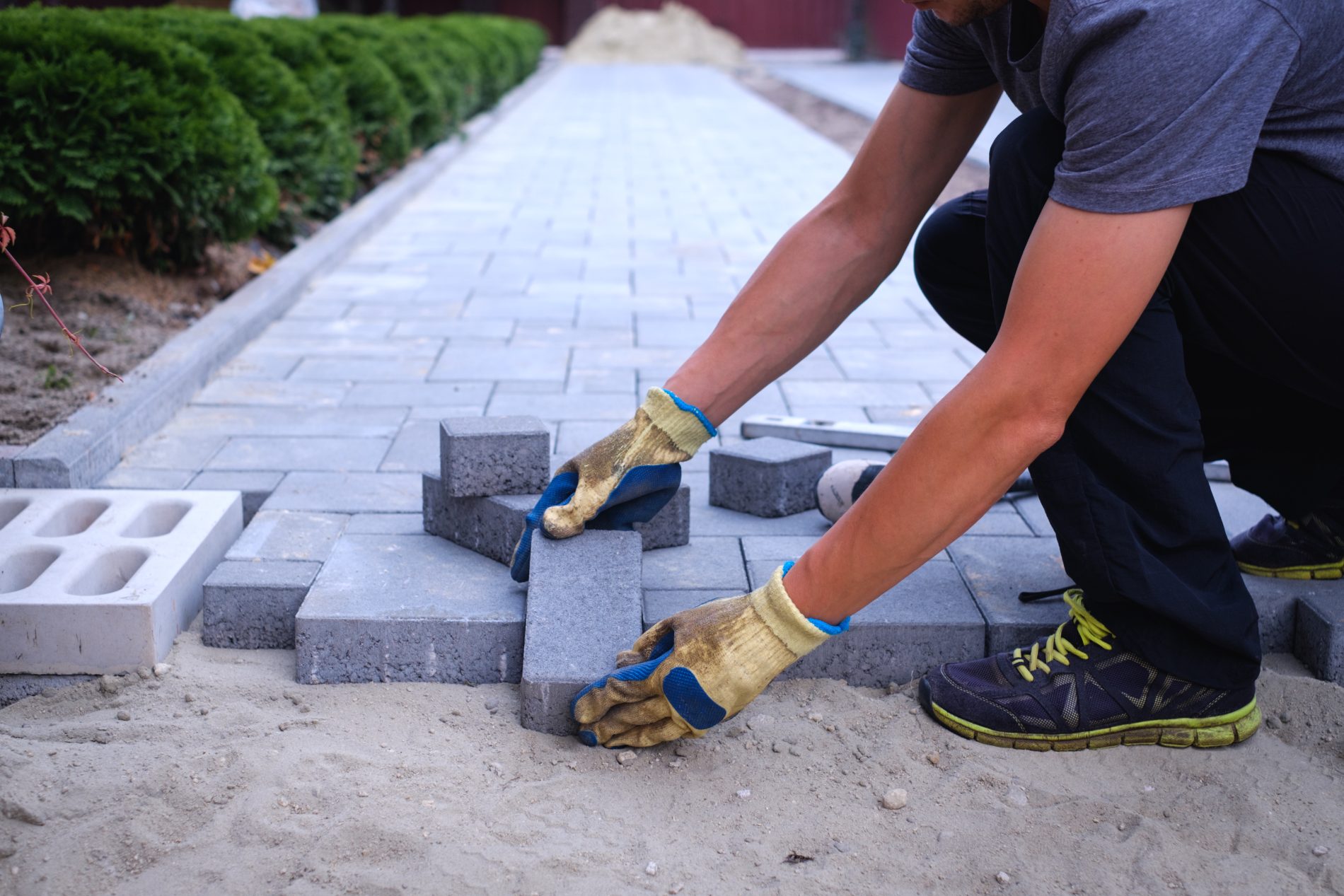 Worker in gloves laying gray concrete pavers on sand base for patio installation